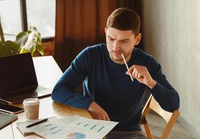 Thoughtful Millennial Guy Looking Through Data Graphs And Charts Thinking On Business Startup Sitting In Coworking Workspace. Selective Focus photo