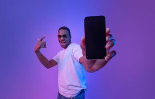 Cool Mobile Offer. Excited African Guy Demonstrating Smartphone With Black Screen, Standing Under Neon Lighting, Joyful Millennial Man Showing Free Mockup Copy Space For Your App Or Website Design photo