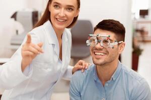 Eye Test Concept. Beautiful lady optometrist checking patient's vision with trial frame, pointing at chart photo
