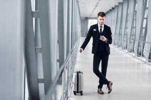 Portrait of confident guy in suit using cell phone standing in airport lobby holding trolley bag, copy space, full length photo