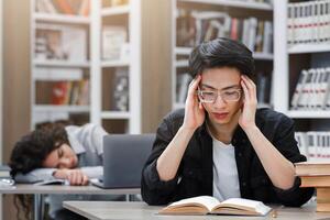 Preparing For Exam Session. Focused asian boy in specs massaging temples, female student sleeping in the background photo