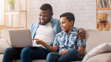 Young black father and his preteen son using laptop at home together, looking at computer screen, making online shopping photo