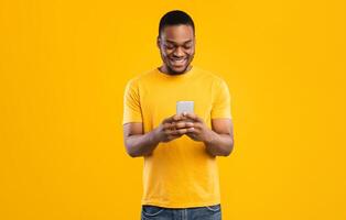 Smiling Black Man Using Smartphone Browsing Internet And Scrolling Social Media News Feed Standing Over Yellow Studio Background. Millennial Guy Using New Mobile Application. People And Gadgets photo