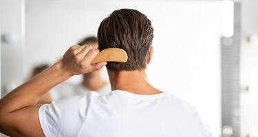 Hair Care. Close up rear view of young man hairbrushing with comb, looking in mirror photo