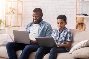 Online Gaming Concept. Happy Afro Father And Son Playing Games On Laptops Together, Sitting With Computer On Laps At Home photo