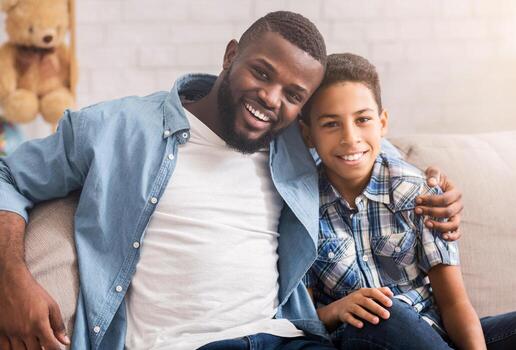 padre y hijo vinculación retrato de contento africano americano preadolescente chico posando a cámara con su papá, abrazando en sofá a hogar foto