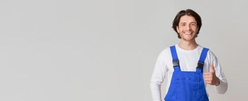 Smiling young laborer posing in work overalls and showing thumb up over light studio background, full-length shot, copy space photo