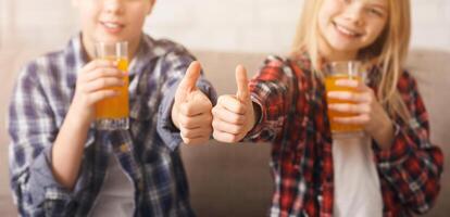 Smiling Boy And Girl Drinking Orange Juice Gesturing Thumbs Up Sitting On Sofa At Home. Shallow Depth, Selective Focus photo