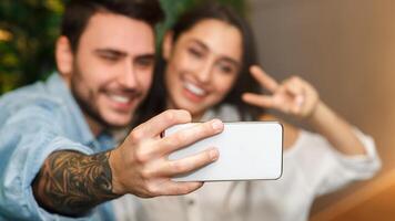Happy Couple Making Selfie On Smartphone Gesturing V-Sign Having Fun During Romantic Date Sitting Together In Cafe. Selective Focus photo