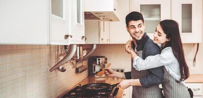 Cooking Together Concept. Lovely couple preparing dinner, frying meat on pan, wife embracing husband from the back photo