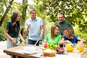 A cheerful group of friends gathers around a table, sharing food and laughter while grilling. The atmosphere is lively, with drinks and a variety of dishes laid out for enjoyment. photo