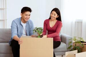 Relocation. Joyful Asian Couple Moving House And Packing Plant In Cardboard Box Sitting On Sofa At Home. Copyspace photo