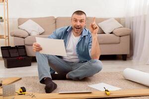 Excited Man Assembling Furniture Pointing Finger Up Having Idea Sitting On Floor At Home, Reading Installation Instruction. Selective Focus photo