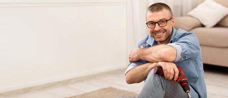Mature Man Assembling Furniture Sitting Posing With Electric Drill, Smiling To Camera At Home. Carpentry Concept photo