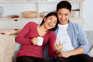 Spending Time Together. Asian family using smart phone sitting on couch at home, empty space photo