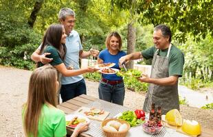 A group of five friends gathers in a vibrant garden to share a meal, smiling and passing around plates of fresh, colorful food during a sunny summer day. photo