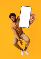 Excited black guy holding smartphone, showing blank screen, jumping up over orange studio background. Handsome african american young man recommending new mobile application, collage photo