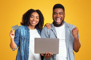 Easy Online Payments. Portrait of joyful african couple celebrating success with laptop and credit card over yellow background photo
