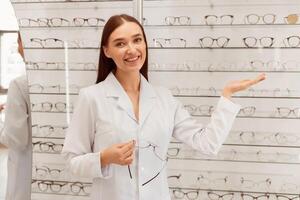 Lady Ophthalmologist. Happy seller showing eyeglasses assortment on the display shelf showcase in optical store photo