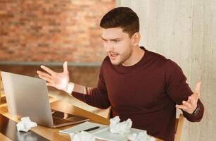 Angry Employee Guy At Laptop Gesturing With Hands Having Problem With Computer Sitting In Modern Office. Selective Focus photo
