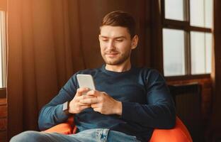 Mobile App. Student Guy Using Phone Texting And Browsing Internet Sitting In Beanbag Chair Indoor. Selective Focus photo