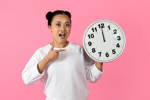 Asian woman smiles widely while holding a round clock and pointing at it, set against a vibrant pink backdrop. The atmosphere is cheerful and lively, emphasizing the time displayed. photo