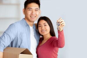Homeowners. Smiling chinese couple showing key to camera, man holding box, selective focus, copy space photo