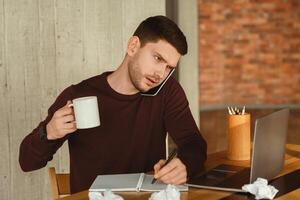 Work Multitasking. Overworked Office Guy At Laptop Talking On Phone Having Coffee And Taking Notes In Modern Office. Selective Focus photo