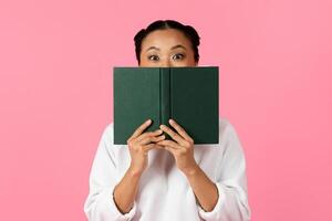 A woman with an excited expression playfully holds a green book in front of her face, against a vibrant pink background, showcasing a moment of joy and surprise. photo