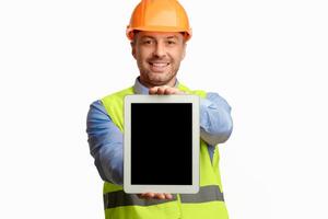 Repairman Showing Digital Tablet Empty Screen Standing Over White Studio Background, Wearing Vest And Helmet. Selective Focus, Mockup photo
