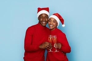 A joyful black couple dressed in matching red sweaters and Santa hats raises glasses of champagne, celebrating the holiday season with smiles against a light blue background. photo