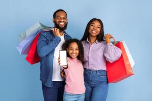 Portrait of happy family of three people of color holding shopping bags, cheerful girl showing smartphone with white empty screen in hand, isolated on blue studio background, mock up, blank space photo