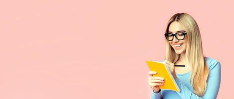 Writing Checklist. Portrait of smiling girl student taking notes in notepad isolated over pink studio wall, copyspace photo