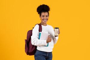 A smiling student holds a takeaway coffee cup and notebooks, wearing a backpack. The vibrant yellow backdrop adds a lively touch to this academic moment. photo