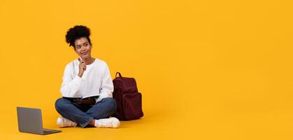 A student sits cross-legged on a bright yellow floor, holding a notebook and pen while looking contemplative. A laptop and a backpack are placed beside her as she engages in study, copy space photo
