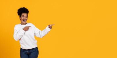 A young woman smiles and joyfully points to copy space, dressed in a white sweatshirt and jeans, set against a vibrant yellow backdrop that enhances the lively atmosphere. photo