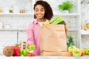Self Isolation Concept. Black girl unpacking paper bag with food in kitchen, ready for quarantine during epidemic outbreak photo