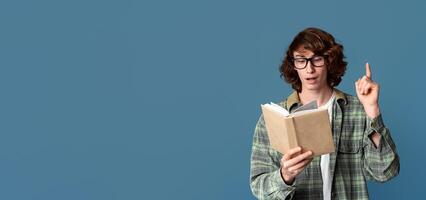 Young hipster guy in glasses reading book and pointing finger up, having great idea, turquoise background photo