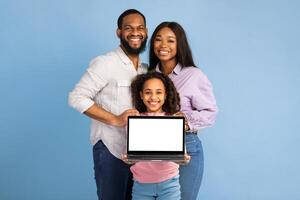 Portrait of happy family of three African American people showing laptop with white empty screen, smiling girl holding gadget with blank space for mock up, standing over blue studio background photo