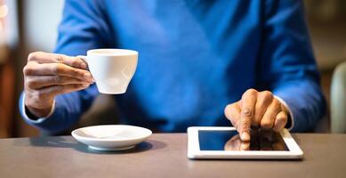 Young afro man sitting at the table with cup of coffee using digital tablet, man's hands typing message on touch screen photo