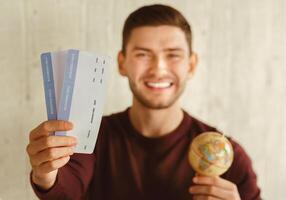 Vacation. Smiling Traveler Guy Showing Flight Tickets Holding World Globe Ready For Journey Sitting Indoor. Selective Focus, Shallow Depth photo