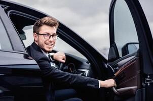 Smiling manager in glasses sitting in luxury car, getting in or out his auto, keeping door open, looking at camera photo