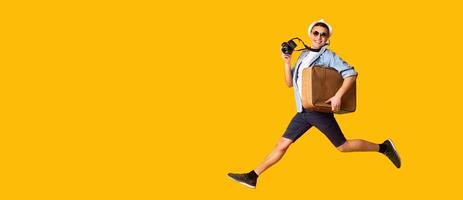 Excited young man with camera and suitcase running towards empty space over yellow background, panorama photo