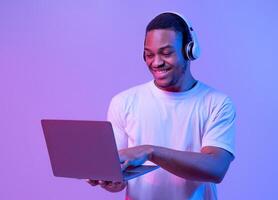 Online Gaming. Portraif Of Happy Black Man In Wireless Headphones Using Laptop Computer, Playing Games In Internet While Standing In Neon Lighting Over Purple Studio Background, Copy Space photo