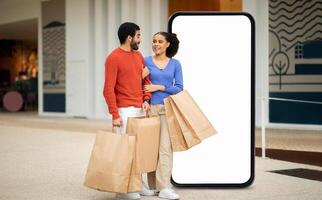 A young couple smiles at each other as they stroll through a shopping mall, holding several paper shopping bags, with a large blank screen behind them showcasing a vibrant shopping atmosphere. photo