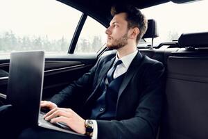 Always In Touch. Handsome businessman working on laptop, sitting in backseat of luxury car, looking out of window photo