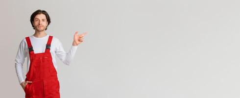 Young confident workman in uniform pointing aside at copy space on light background, panorama photo