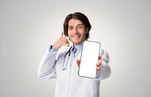 Positive medical specialist pointing at camera and showing call gesture over white studio background, empty space photo