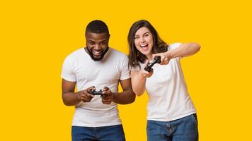 Joyful interracial couple holding joysticks competing with each other in games, standing together over yellow background, empty space photo