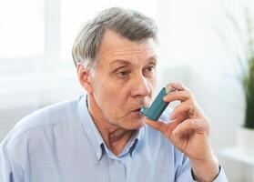 Senior Man Using Asthma Inhaler For Allergies In Modern Clinic, Copy Space photo